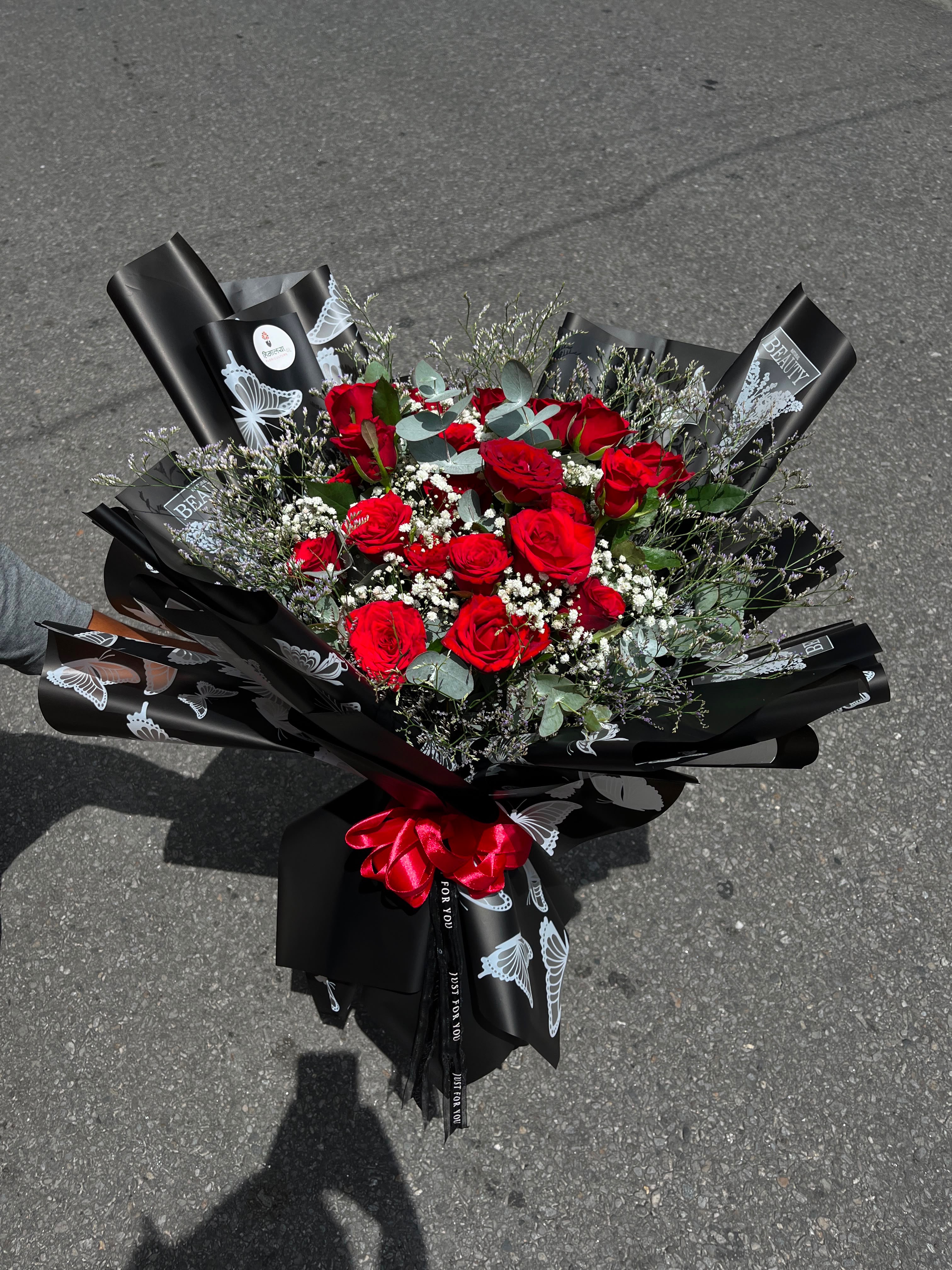 Grand Red Rose Arrangement A bold bouquet featuring 20 stems of red roses arranged in a bulky design, complemented by dollar leaves and gypsophila, with elegant black wrapping for a dramatic finish.
