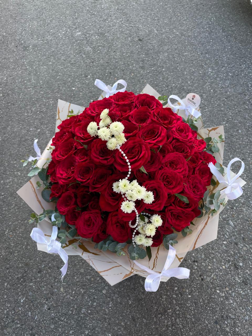 A spectacular premium bouquet featuring 100 fresh red roses, beautifully arranged in a full and voluminous design with elegant Dollar Leaves, delicate white daisies, and pearl accents. The arrangement is finished with a soft baby pink wrapper and a white bow ribbon, creating a luxurious and romantic presentation.