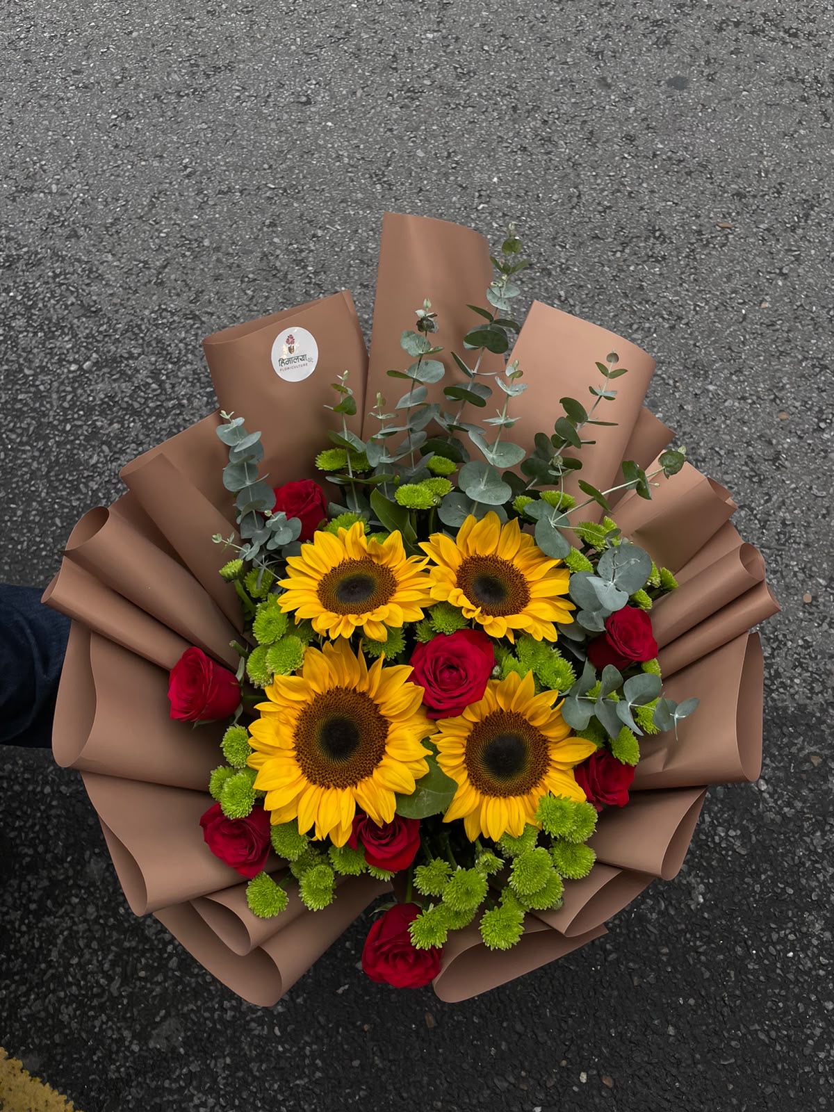A vibrant mixed floral arrangement featuring 8 red roses and 4 sunflowers, complemented with green daisies and Dollar Leaves. The flowers are arranged on an Oasis base with a full, bulky design and finished with an elegant brown wrapper.