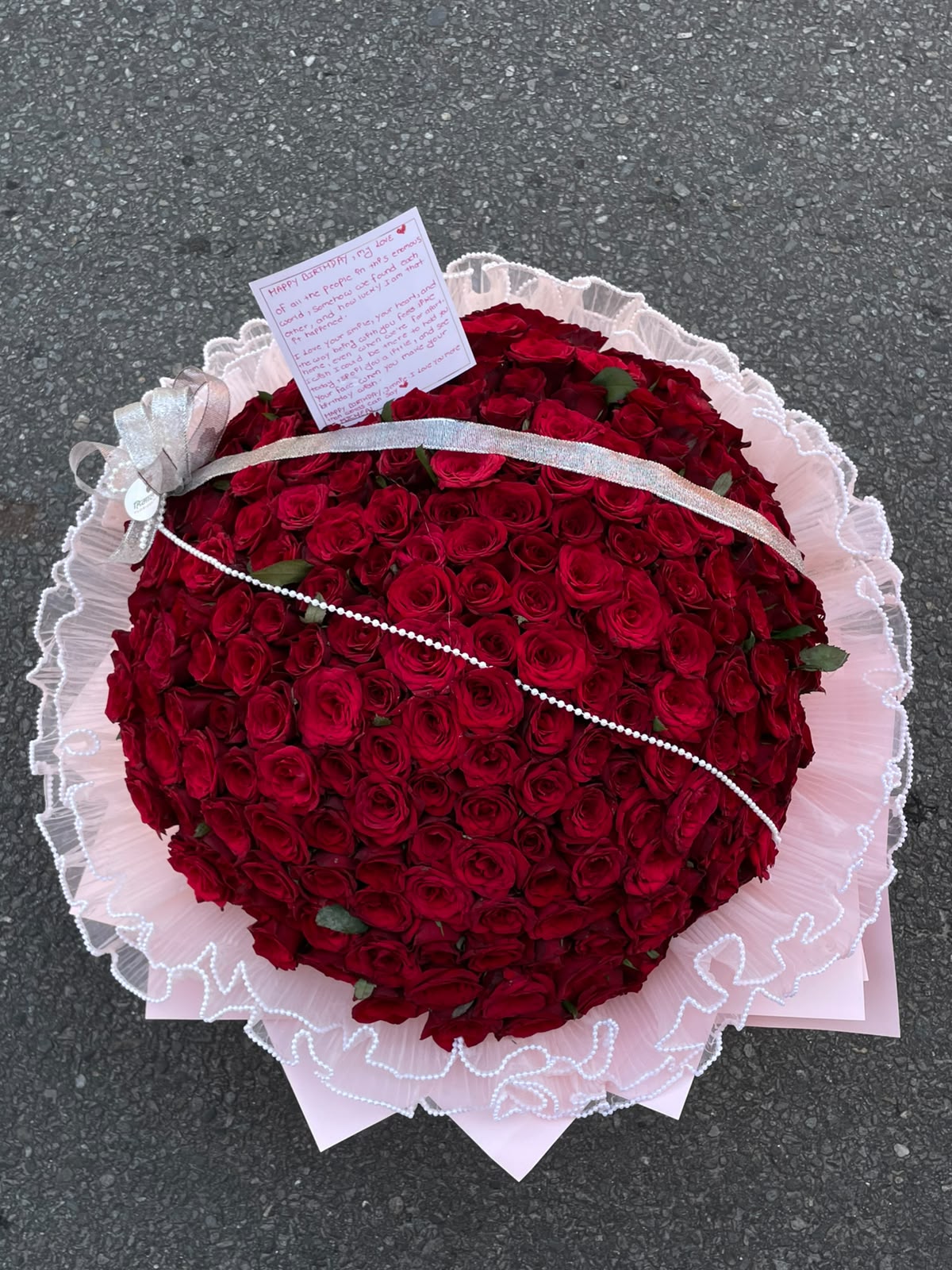 A magnificent floral arrangement featuring 200 red roses, beautifully presented in a baby-pink wrapper with decorative pearl detailing. The combination of rich red blooms and soft pink accents creates a grand and romantic presentation.