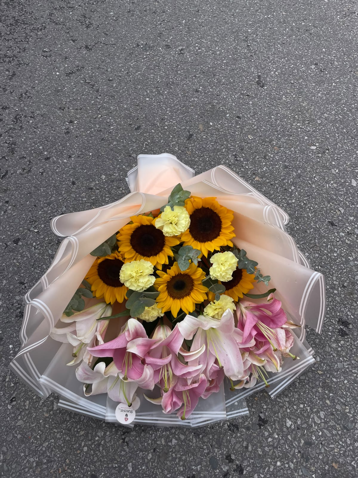 Sunshine Lily Bouquet A beautiful mixed floral arrangement featuring 5 sunflowers, 4 yellow carnations, and 4 stems of pink lilies, complemented with Dollar Leaves and elegantly finished with a white wrapper. The combination creates a bright, fresh, and colorful presentation.