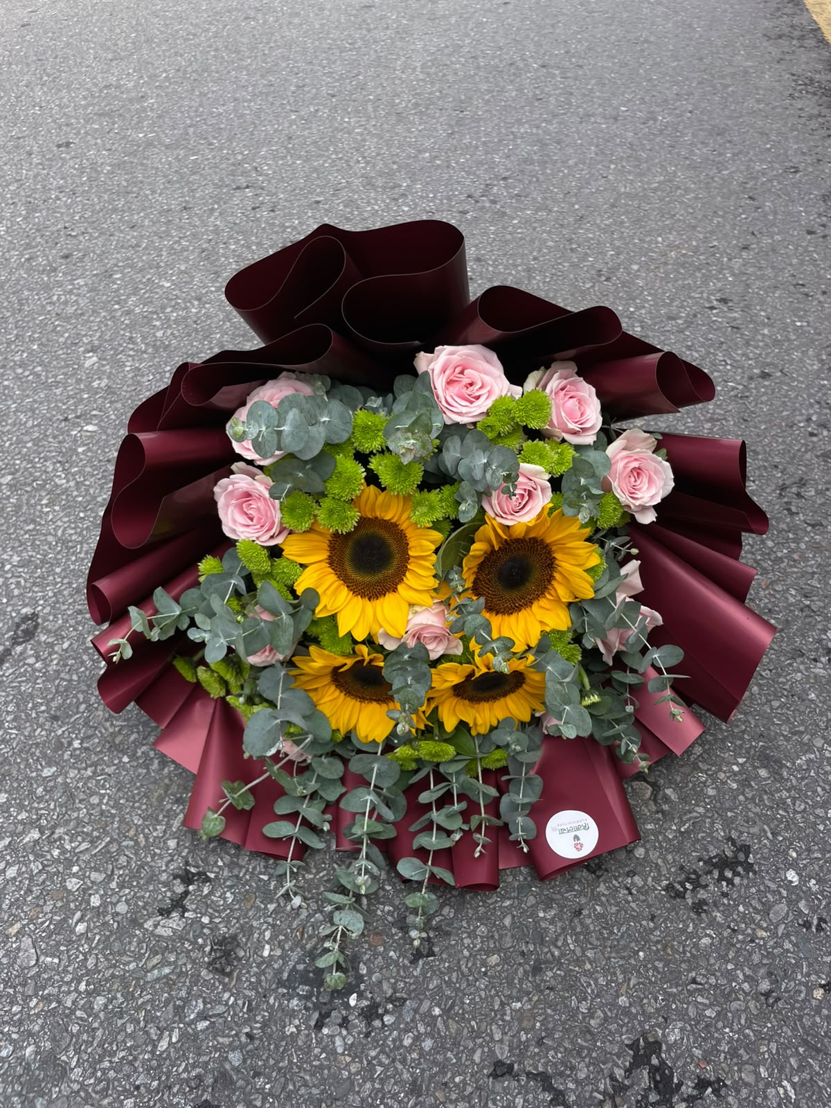 Maroon Sunshine Bouquet A vibrant mixed bouquet featuring 4 sunflowers and 12 stems of pastel roses, complemented by Dollar Leaves and Green Daisies. The arrangement is beautifully finished with a premium maroon wrapper, creating a rich and elegant contrast.
