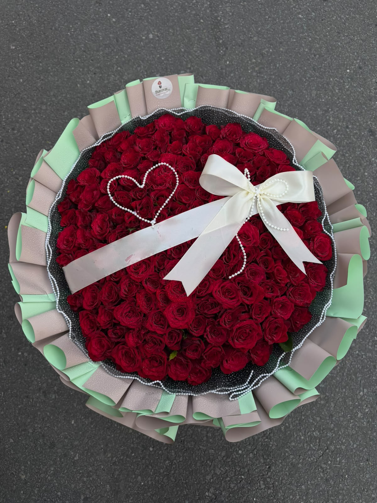 A grand floral arrangement featuring 150 red roses in a bulky design, styled with pearl wrapping, light green and light pink paper, and a white ribbon. The oasis base provides support for this impressive statement bouquet.