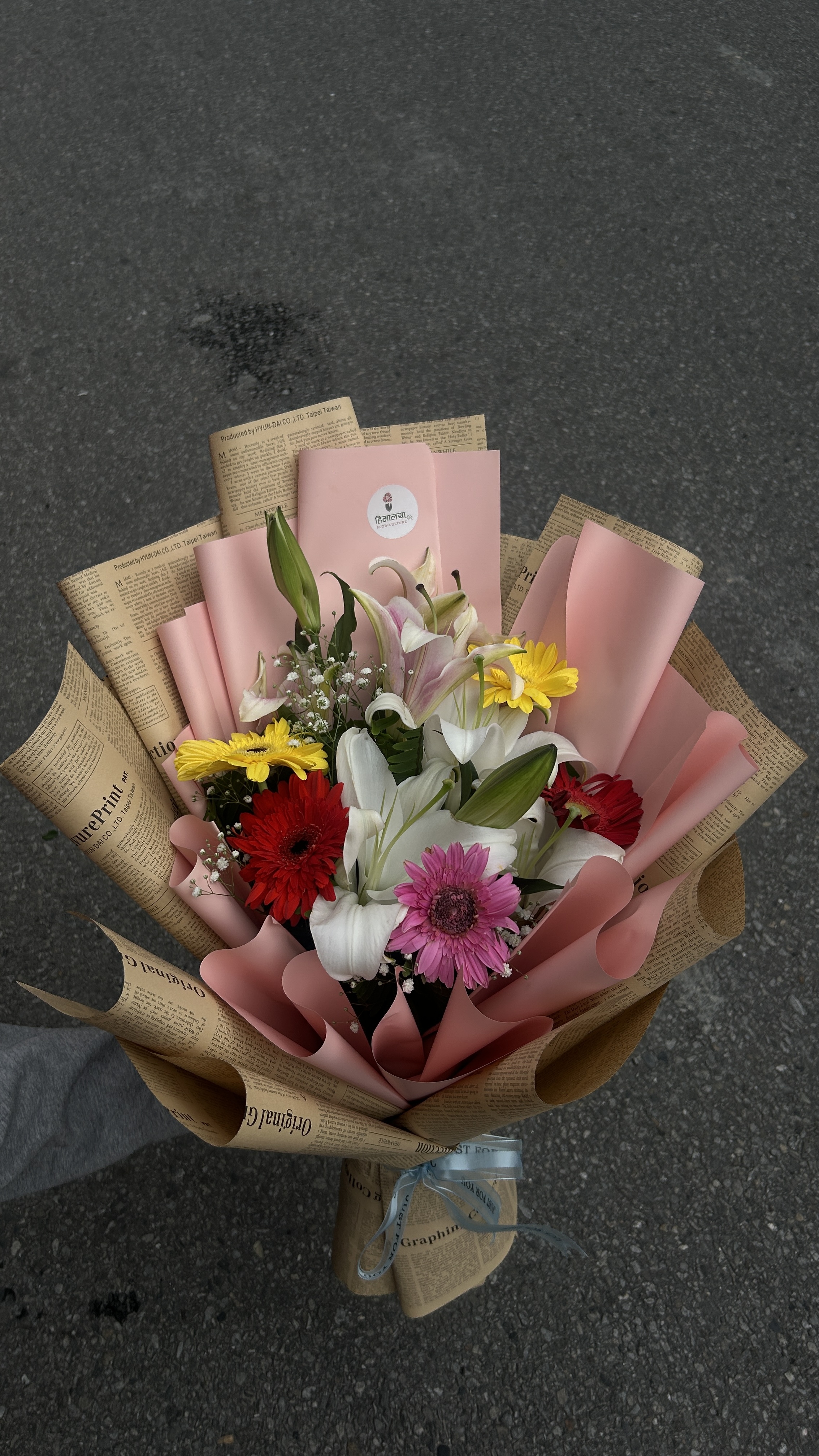 A beautiful combination of pink and white lilies with five mixed-color gerberas, complemented by delicate Gypsophila. The pastel pink and premium newspaper wrapping create a soft, elegant, and stylish presentation.