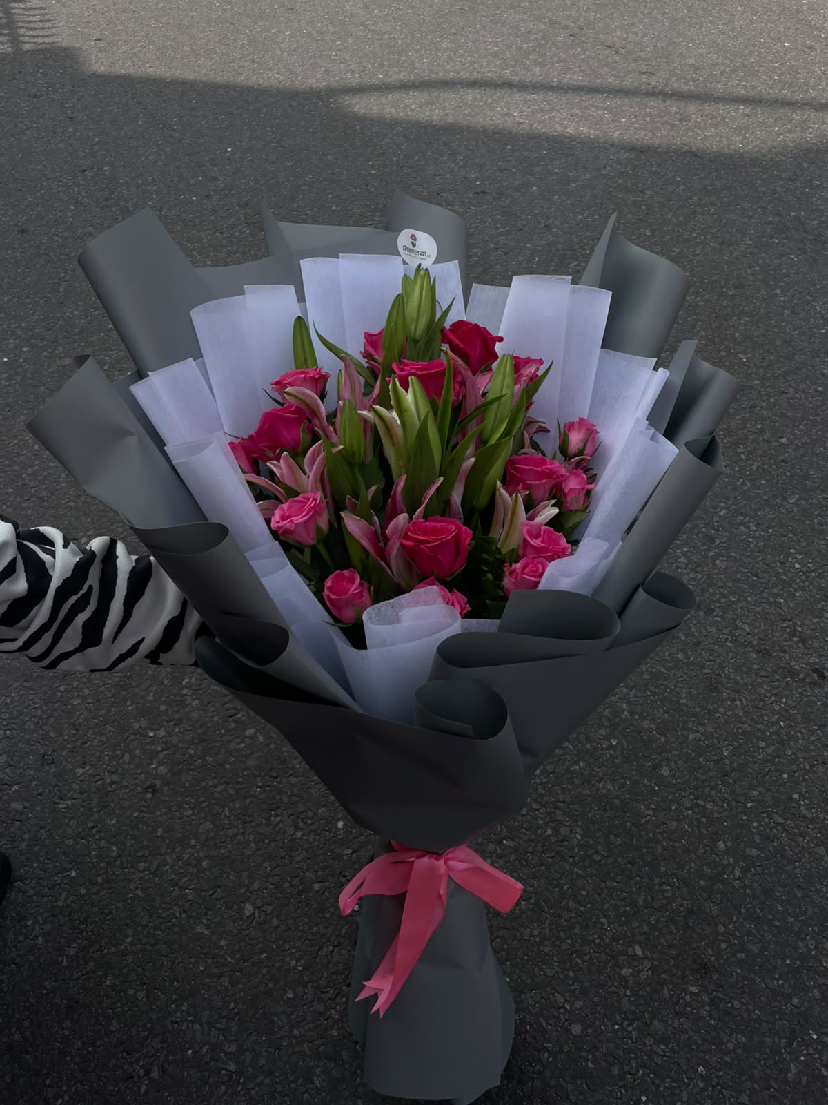 A striking floral arrangement featuring four pink lilies and 20 hot pink roses, finished with a white and grey wrapper and a pink ribbon for a bold yet elegant presentation.